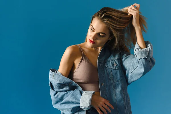 Un modelo de moda en chaqueta de mezclilla, top beige y jeans se apoya sobre un fondo azul. Hipster mujer urbana en el estudio. Moda americana — Foto de Stock