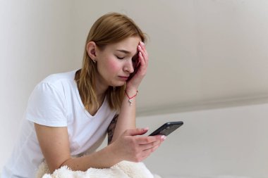 Depresyonda olan genç ve güzel bir kadının portresi. Yatakta oturmuş, telefonunu tutuyor. Siber zorbalık, akıllı telefon bağımlılığı ve depresyon, ayrılık, üzücü haber.