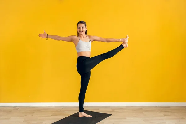 Ung kvinna stående i yoga asana utthita hasta Padangusthasana, förlängd hand till stortå yoga pose, studio skott, gul bakgrund — Stockfoto