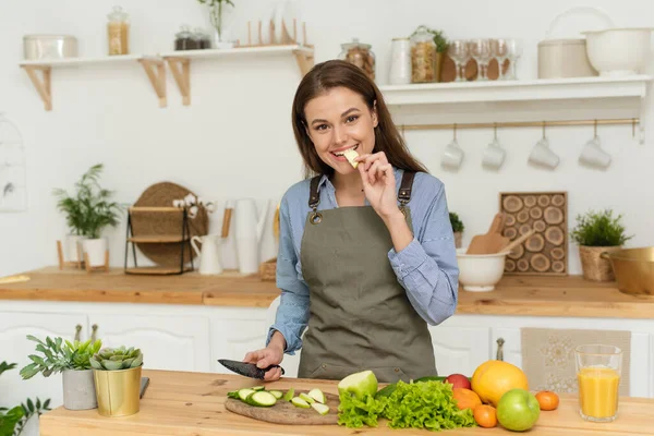 Mooie vrouw die ontbijt klaarmaakt in een gezellige houten keuken. Een jonge vrouw neemt een hap van een appel en kijkt in de camera — Stockfoto