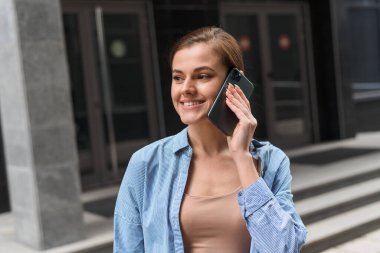 Bir iş kadını bir ofis merkezinin arka planında yürüyor ve telefonda konuşuyor. İş ortaklarıyla uzaktan iletişim