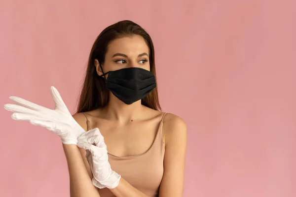 Närbild foto av ung kvinna i en medicinsk mask sätter på skyddande kirurgisk sterila handskar på armen på rosa bakgrund, skydd mot coronavirus — Stockfoto