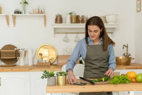 Gezond eten thuis. Gelukkige jonge vrouw met behulp van de telefoon het bereiden van groente salade in de keuken — Stockfoto