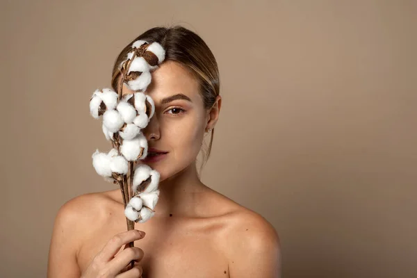 Retrato de mujer joven y bonita con rama de algodón suave con piel limpia y fresca en estudio sobre fondo beige. Cosmetología, belleza y concepto de spa. — Foto de Stock