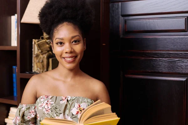 Retrato de uma jovem estudante negra. Ela está de pé na biblioteca da universidade, segurando um livro e olhando para a câmera — Fotografia de Stock