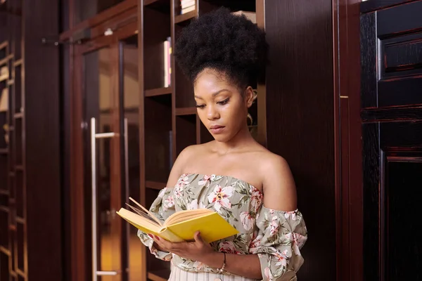 Jovem africana compra livros em uma livraria, lendo e escolhendo-os. modelo encantador lê um livro sobre o fundo de armários de madeira — Fotografia de Stock