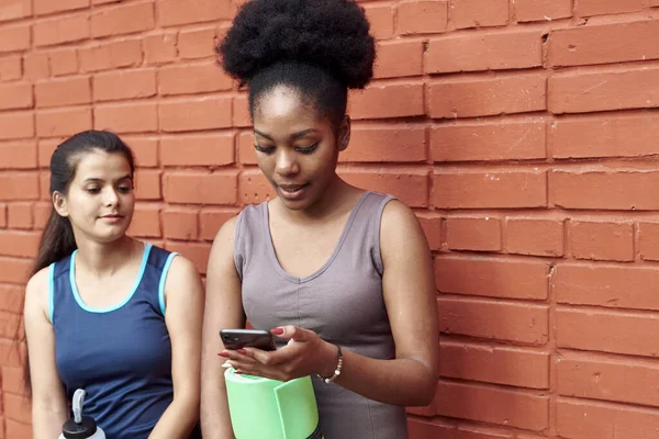 Elképesztő fiatal atlétikai nők képe egy téglafal előtt, ahogy belenéznek a telefonba. Két női barát nevet a közösségi média poszton. — Stock Fotó