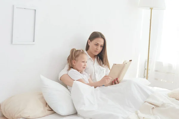 Close-up mom and little daughter are resting in a cozy bed, reading a funny interesting childrens book together — Stock Photo, Image