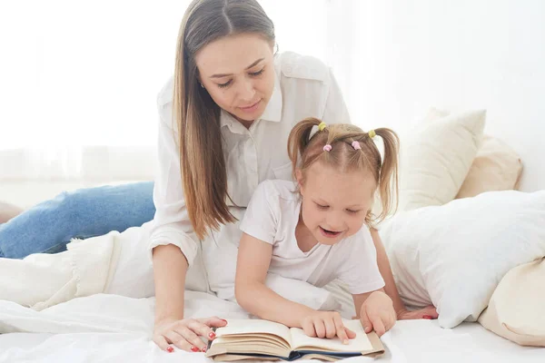 Mosolygó fiatal anya vagy dadus ül az ágyban aranyos kislány gyermek olvasás érdekes könyvet, töltik a hétvégét a hálószobában együtt — Stock Fotó