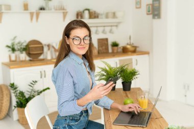 Genç bir kadın mutfak masasında oturuyor, işe ara veriyor, telefonda sosyal ağlarda oturuyor ve kameraya bakıyor. Genç, güzel, serbest çalışan bir bayan.