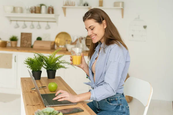 Feliz bela jovem sentada à mesa em casa, trabalhando em um laptop e bebendo suco de laranja delicioso. Trabalho e estudo em casa em condições de quarentena — Fotografia de Stock