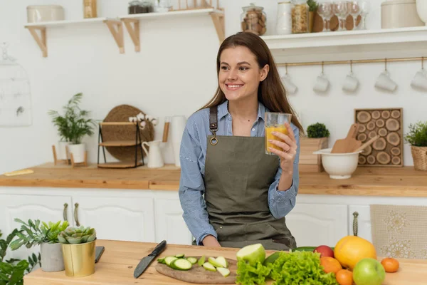 Mulher bonita preparando café da manhã em uma cozinha de madeira acolhedora. Uma jovem está segurando suco de laranja e está de pé em sua cozinha brilhante — Fotografia de Stock