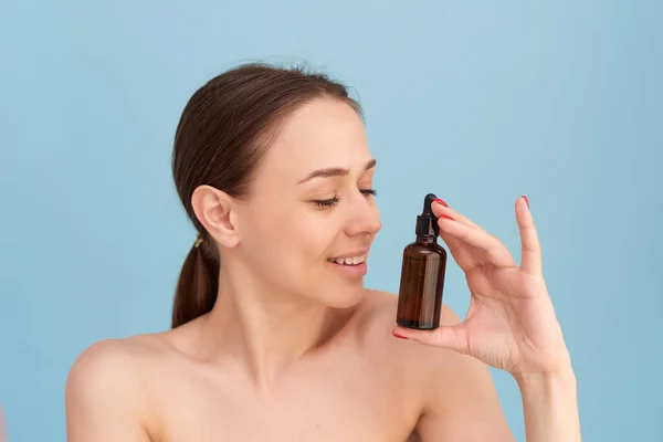 Smiling woman holding vitamin c serum near her face on blue background. Skin care and health concept — Stock Photo, Image