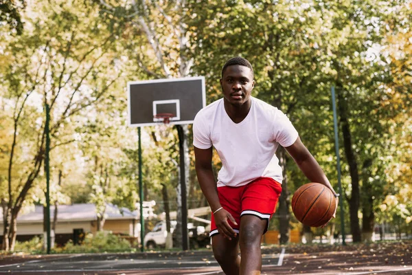 Young black smiling man playing basketball on the court at sunrise, morning sports. Outdoor portrait
