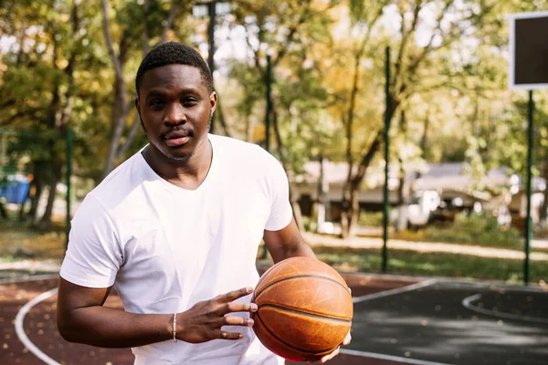 Um jovem afro-americano está jogando basquete em uma quadra ao ar livre, sorrindo e olhando para a câmera. Esportes ao ar livre — Fotografia de Stock