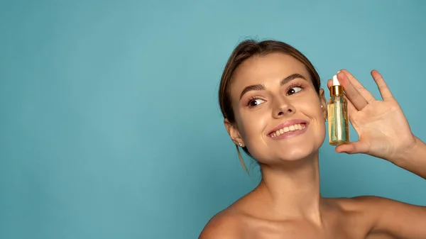 Mujer sonriente sosteniendo suero de vitamina C cerca de su cara sobre fondo azul. Cuidado de la piel y concepto de salud. Banner web — Foto de Stock