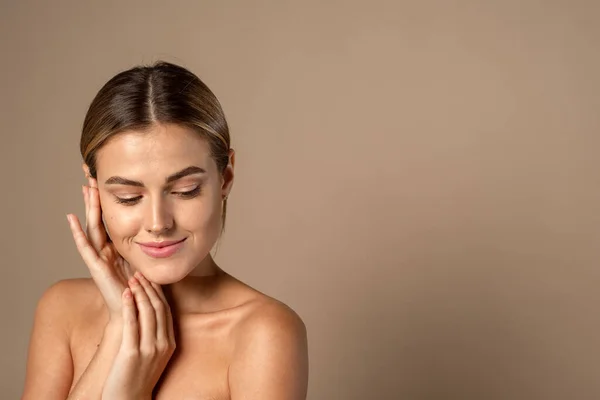 Cuidado de la piel. Mujer con cara de belleza tocando piel facial sana. Hermosa sonriente modelo femenino caucásico con maquillaje natural tocando piel hidratada brillante sobre fondo beige — Foto de Stock