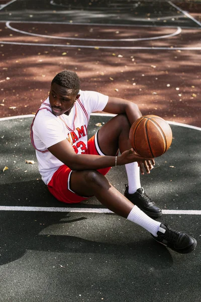 Portré egy afro-amerikai fiatalemberről, aki kosárlabdát tart és egy kosárpályán ül. Tarts szünetet edzés közben. Divatos sport portré — Stock Fotó