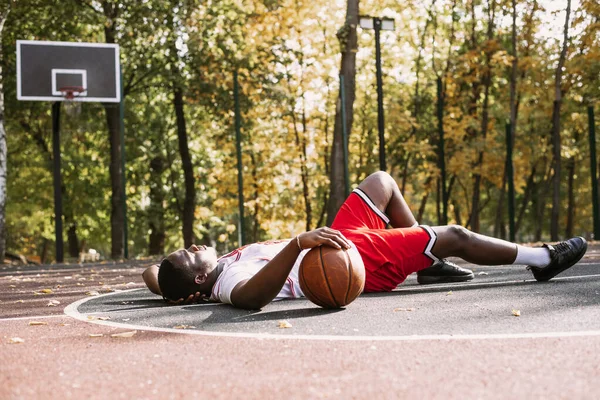 Retrato de um jovem afro-americano segurando um basquete e sentado em uma quadra de basquete. Faça uma pausa durante o treino. Retrato de esportes na moda — Fotografia de Stock