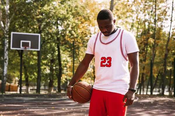 Retrato de um jovem negro bonito segurando uma bola de basquete em um campo de basquete. Faça uma pausa durante o treino. Retrato de esportes na moda — Fotografia de Stock