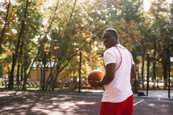 Egy fiatal afro-amerikai férfi kosárlabdázik a szabadtéri pályán, mosolyog és a kamerába néz. Szabadtéri sportok — Stock Fotó