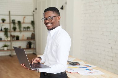 Modern bir ofiste çalışan gülümseyen Afrikalı bir adam dizüstü bilgisayarın arkasında kameraya bakıyor. Gömlekli ve pantalonlu yakışıklı Afro-Amerikan işadamı dizüstü bilgisayar kullanıyor ve çalışırken not alıyor.