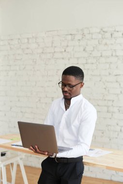 Konsantre olmuş Afrikalı Amerikalı adam modern bir ofiste dizüstü bilgisayar, not defteri ve iş evrakları kullanıyor. Genç siyahi erkek serbest çalışan birlikte çalışma alanında duruyor ve çalışıyor.