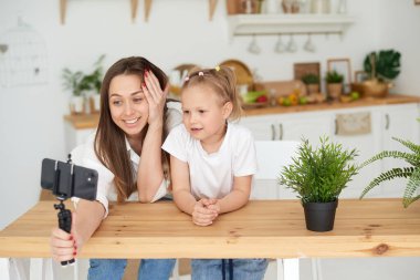 Mutlu bir aile. Sevimli küçük kız mutfakta annesinin yanında oturuyor, gülüyor, akıllı telefon kullanarak eğleniyor, komik bir video izliyor. Karantina sırasında video iletişimi, iletişim