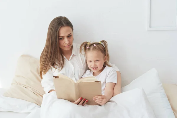 A fiatal, gyönyörű anya könyvet olvas a lányának. Boldog család, anya és lánya olvasás egy könyvet, miközben fekszik az ágyban, mosolygós dada anya mondja vicces történet aranyos óvodás lány — Stock Fotó