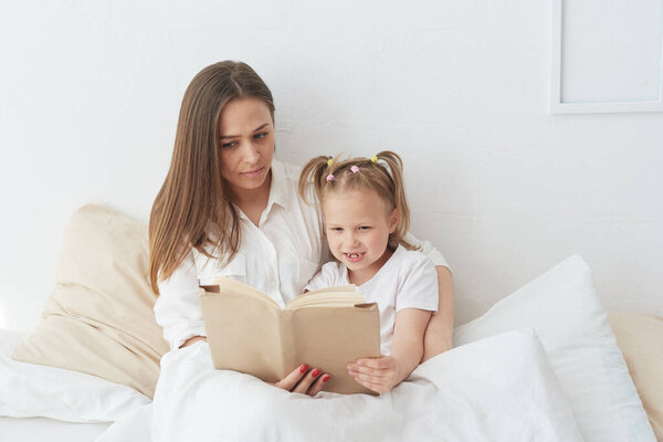 Young beautiful mother is reading a book to her daughter. Happy family, mother and daughter reading a book while lying in bed, smiling nanny mom telling funny story to cute preschool girl