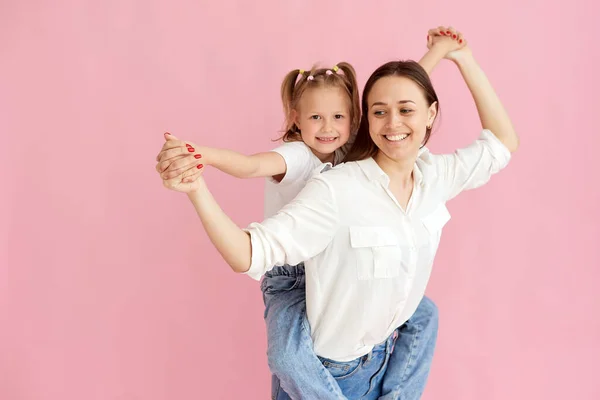 Mama kobieta w lekkich ubraniach bawi się z cute baby girl. Mała córeczka siedząca na plecach samotna na pastelowym różowym tle. portret pracowni — Zdjęcie stockowe