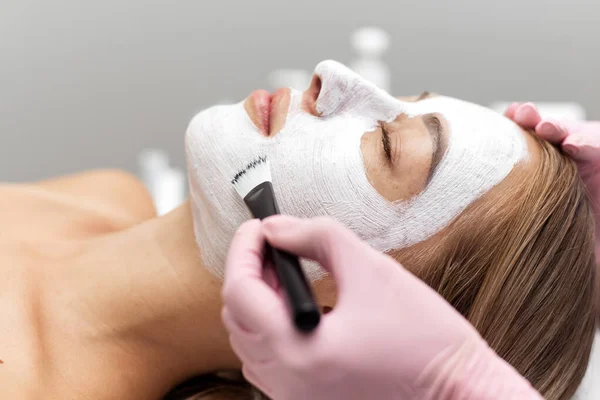 Schoonheidsspecialiste brengt een wit klei gezichtsmasker met een borstel aan op een vrouwengezicht. Professionele schoonheidsspecialiste brengt witte cosmetische crème aan op een meisje dat in een moderne spa op de bank ligt. Sluit maar af. Ontspannen vrouw — Stockfoto