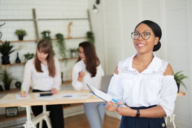 Gülümseyen esmer tenli genç bir kadın, arka planda meslektaşlarıyla modern bir ofiste duruyor. Konferans odasında çalışan kadın tasarımcı.