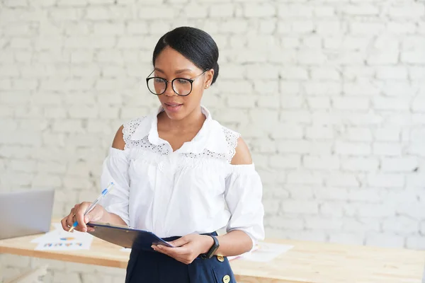 Молодая афроамериканка стоит в стильном офисе и смотрит в блокнот. Самка Миллениал занимается бизнесом или фрилансом — стоковое фото