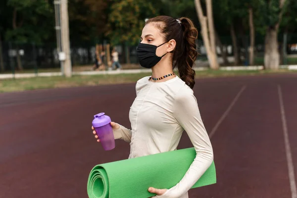 Een jonge vrouw met een zwart beschermend masker staat op het sportterrein en houdt een yogamat en een fles water vast. Sportactiviteiten tijdens quarantaine. Buiten — Stockfoto