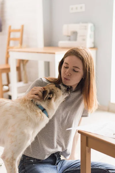 家で犬と抱き合って笑っている赤毛の女性。家にモングレルを避難させろ。ライフスタイル — ストック写真