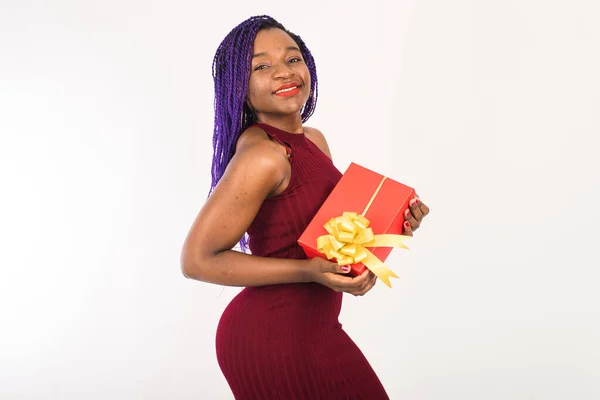 Uma bela mulher afro-americana segura um grande presente vermelho em suas mãos, sorri e olha para a câmera. Isolado sobre fundo branco — Fotografia de Stock