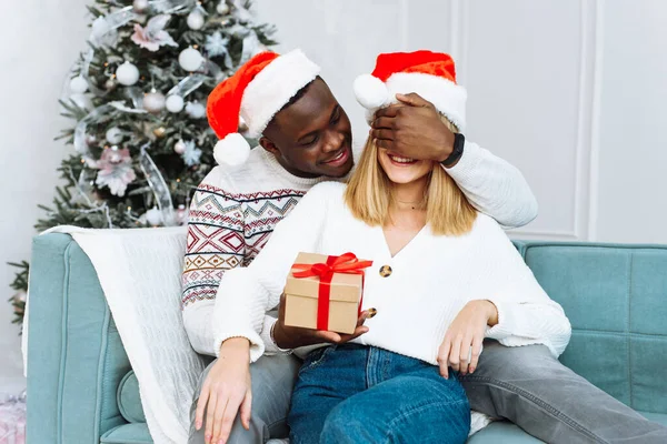 Una joven con los ojos cerrados recibió un regalo de una persona encantadora que la abraza. Concepto de Navidad y Año Nuevo. Pareja romántica intercambiando regalos de Navidad en casa —  Fotos de Stock
