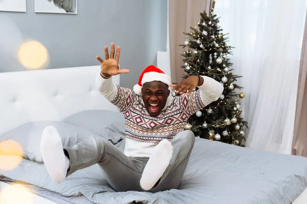 Egy gyönyörű fekete férfi télapó kalapban ül az ágyon a háta mögött egy karácsonyfa, kiabál, felemeli a karját és a lábát. Egy vidám ember élvezi az ünnepeket. Ünnep, karácsony és új — Stock Fotó