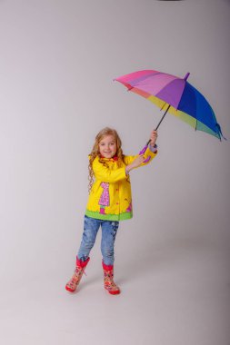 Stüdyoda sarı yağmurlukla gökkuşağı şemsiyesiyle poz veren sevimli küçük kız. 