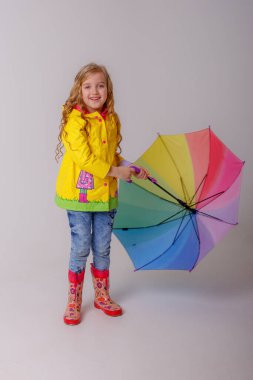 Stüdyoda sarı yağmurlukla gökkuşağı şemsiyesiyle poz veren sevimli küçük kız. 