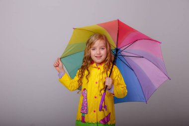 Stüdyoda sarı yağmurlukla gökkuşağı şemsiyesiyle poz veren sevimli küçük kız. 