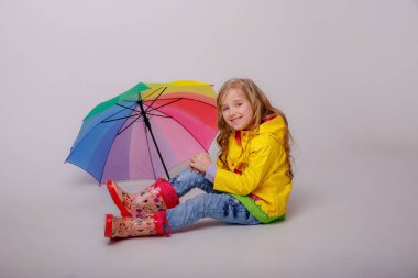 Stüdyoda sarı yağmurlukla gökkuşağı şemsiyesiyle poz veren sevimli küçük kız. 