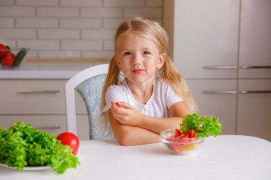 Sevimli sarışın çocuk mutfakta oturmuş sebze yiyor.