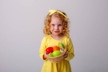 Küçük kıvırcık saçlı bir kız, gri arka planda Paskalya yumurtaları olan sepeti tutuyor.