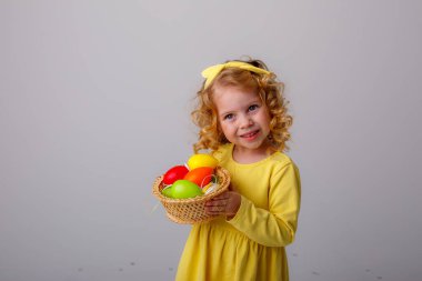 Küçük kıvırcık saçlı bir kız, gri arka planda Paskalya yumurtaları olan sepeti tutuyor.