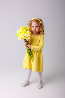 Sarı elbiseli küçük kıvırcık saçlı bir kız elinde gri arka planda bir buket bahar çiçeği tutuyor. 