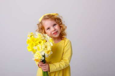 Sarı elbiseli, küçük kıvırcık saçlı bir kız elinde bir buket bahar çiçeği tutuyor.  