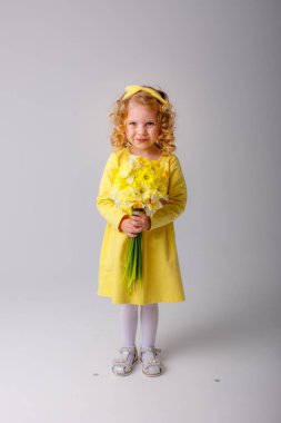Sarı elbiseli küçük kıvırcık saçlı bir kız elinde gri arka planda bir buket bahar çiçeği tutuyor. 