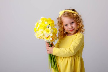 Sarı elbiseli küçük kıvırcık saçlı bir kız elinde gri arka planda bir buket bahar çiçeği tutuyor. 